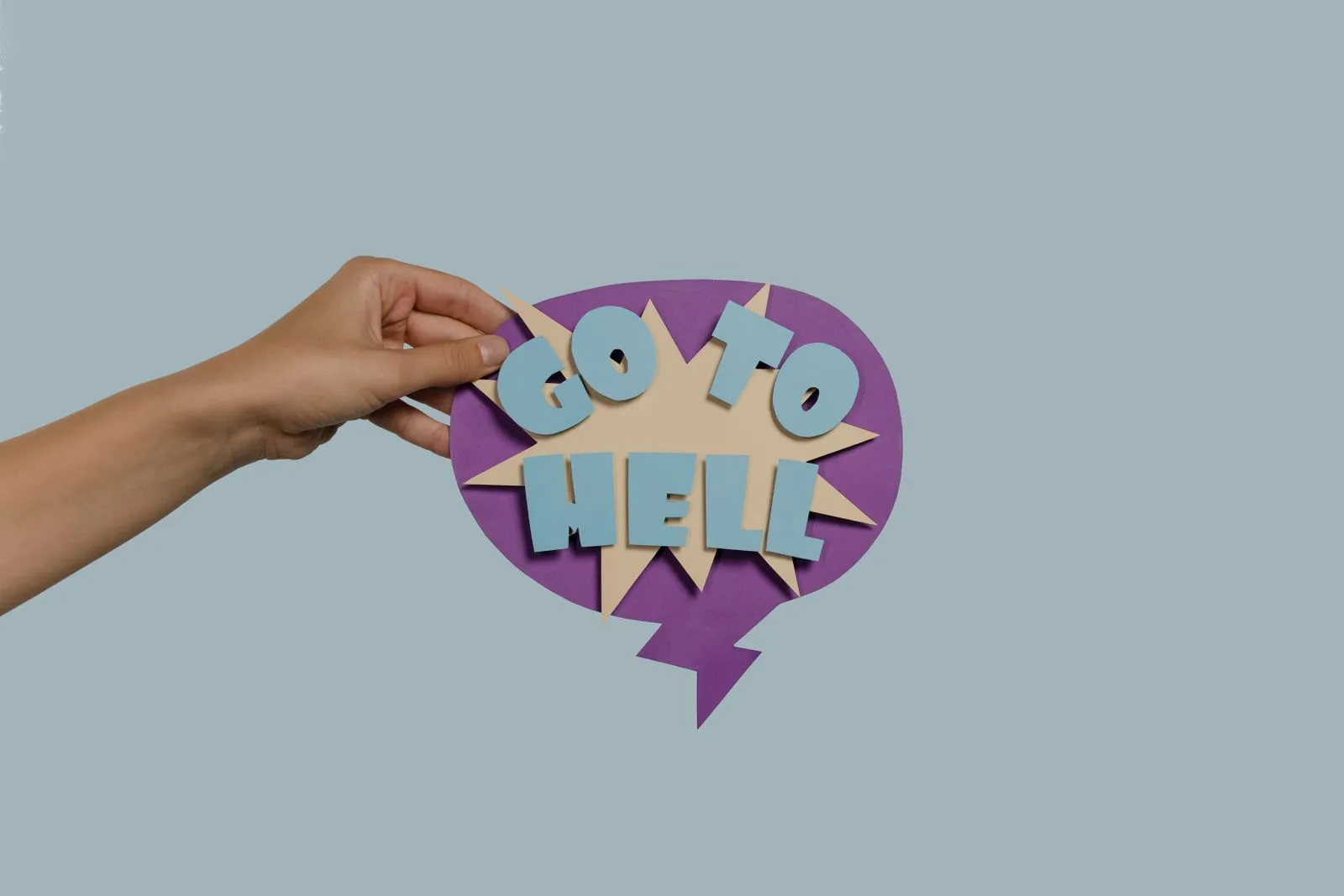 A hand holds a colorful paper cutout with the words 'Go To Hell' on a blue background.